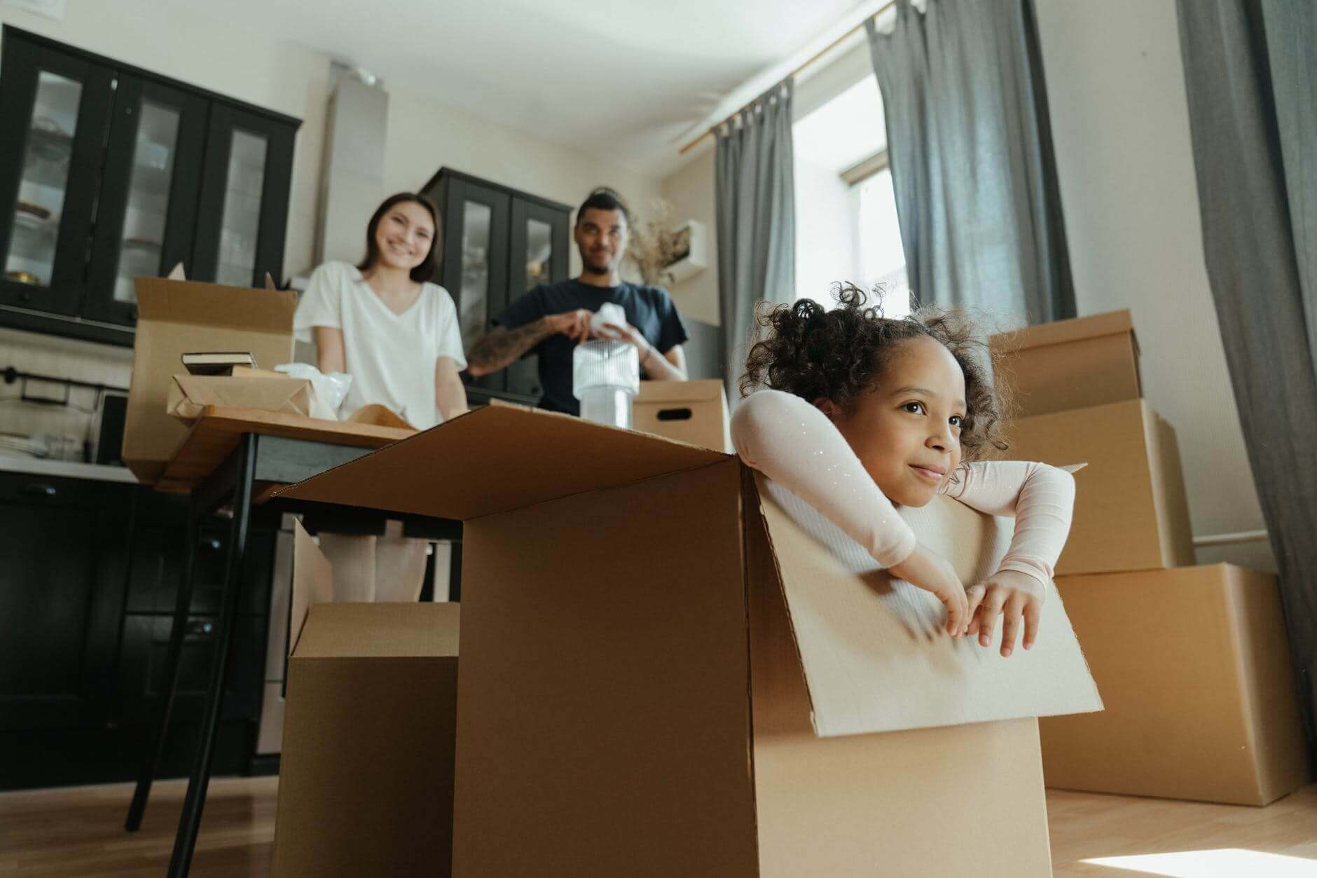 Photograph of a smiling child emerging from a cardboard box. There are two adults behind her, probably the parents, smiling at her. There are lots of other cardboard boxes in the room waiting to be packed.