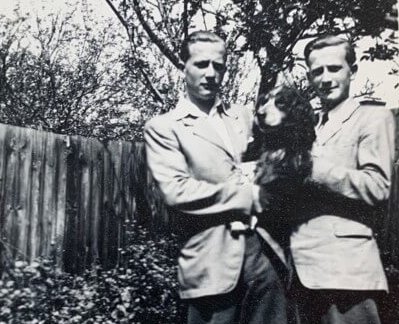 Black & white photograph from the 1950s of 2 smartly dressed young men. They are stood in front of a fence, holding a dog, with a tree behind them. The men are George Coleman's father and his twin brother.