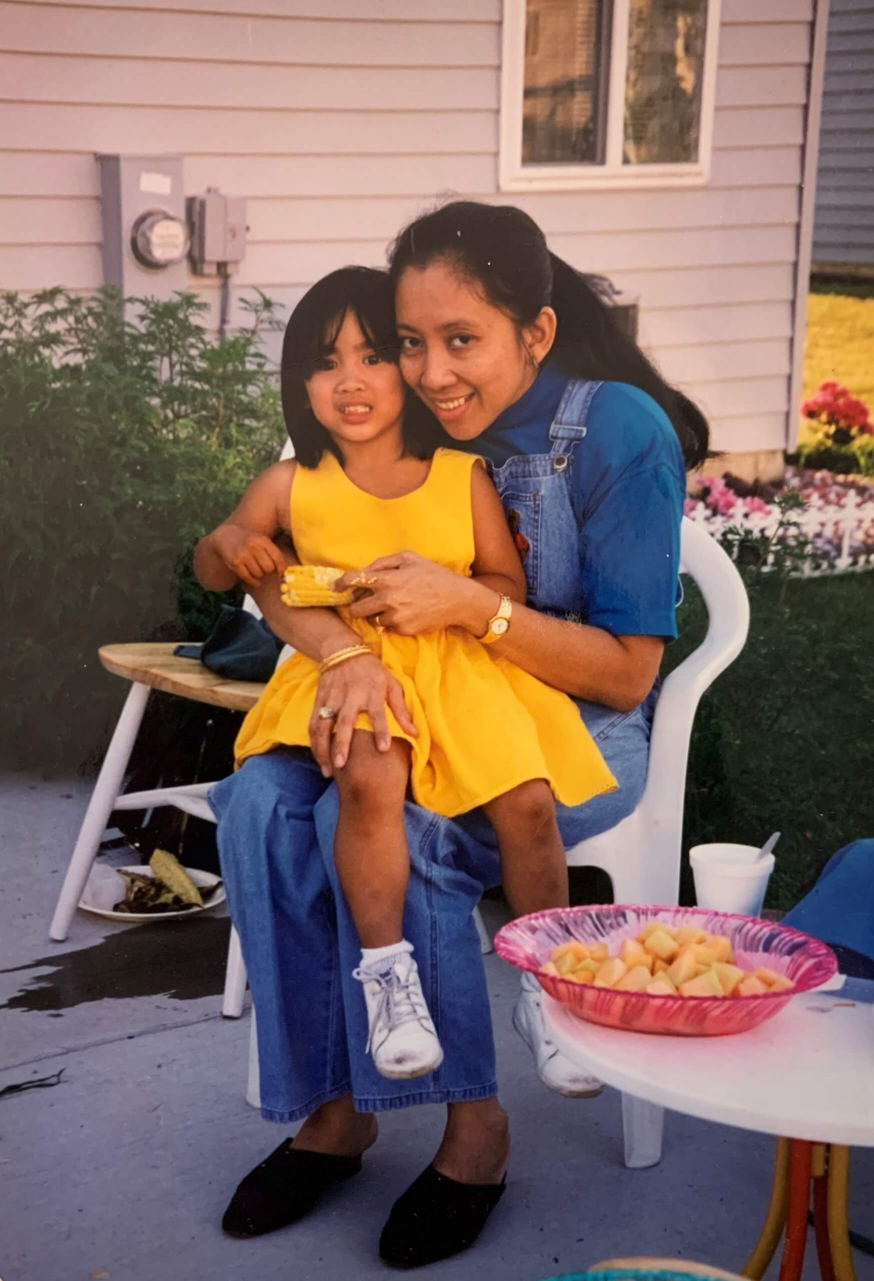 Photograph of a mother and young daughter sitting cuddling on a chair. The mother is on the right, the little girl on the left. The girl is wearing a yellow dress. They look happy.