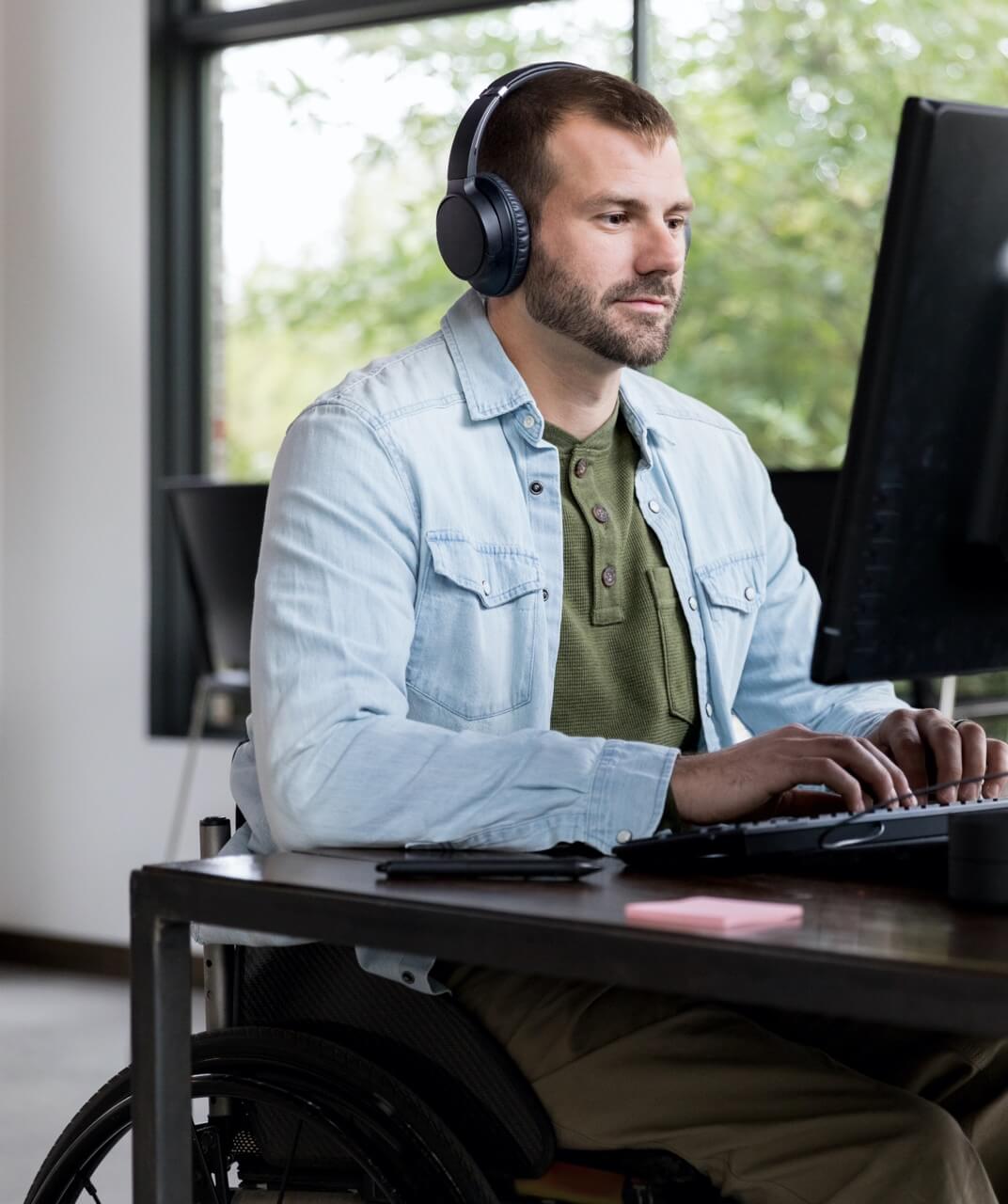 Man in a wheelchair sat working at his computer. He has a beard and is wearing headphones. He is typing at the keyboard and appears to be working in an office.