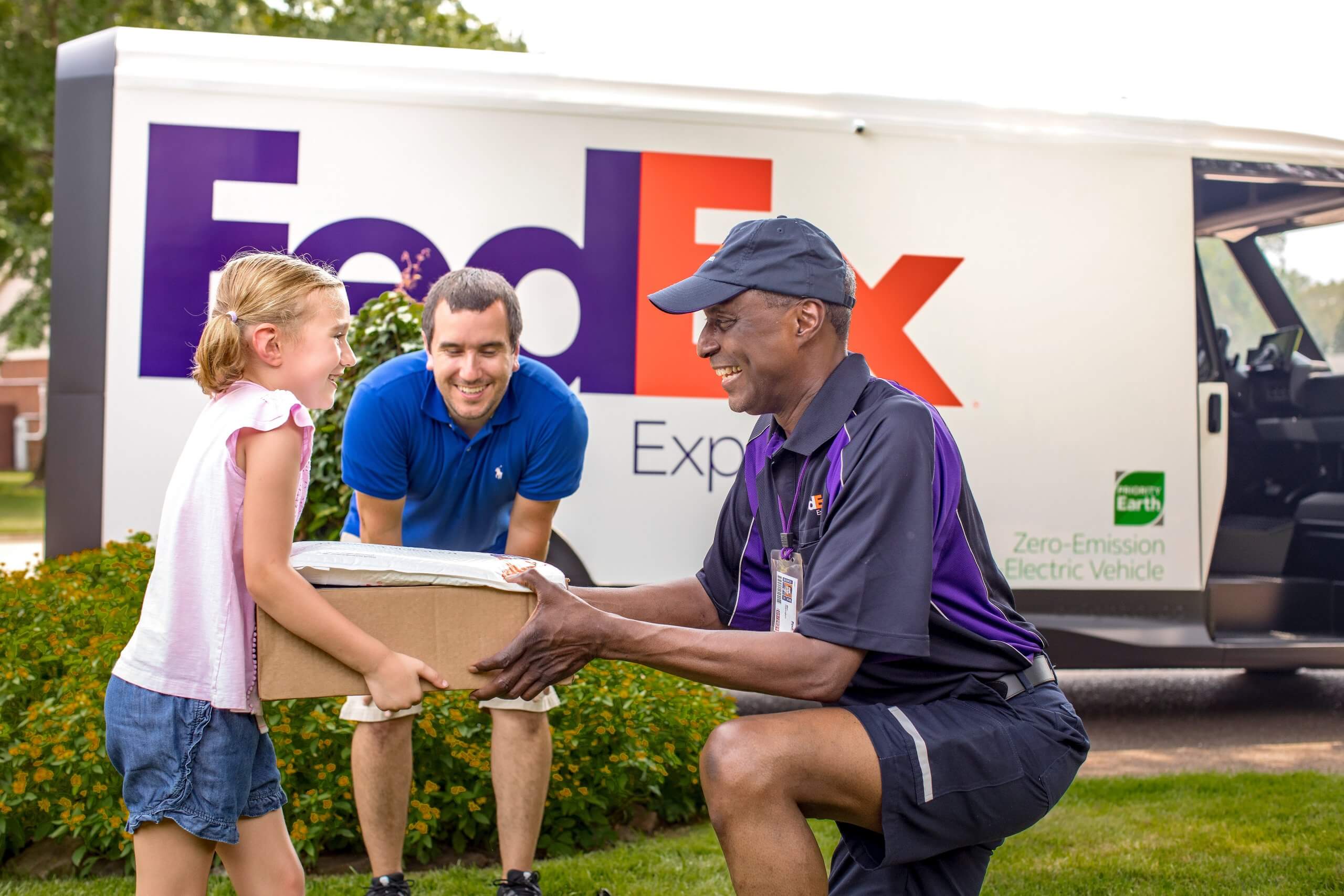 A Fedex delivery man on the right, ,handing a parcel to a girl on the left and her father. There is a Fedex van in the rear of the shot.