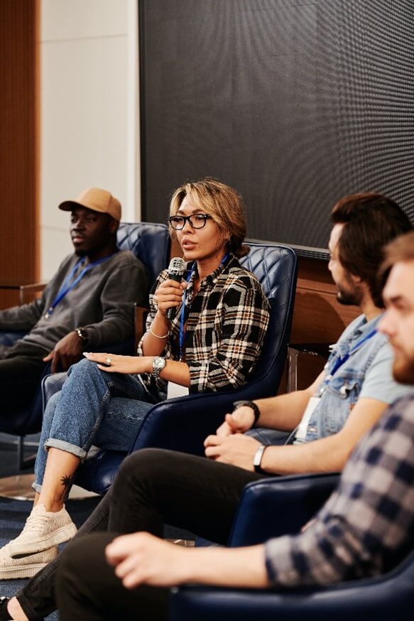 Image of one woman and three men sitting in chairs and talking to a non-pictured audience. Woman is holding microphone and talking while others in the photo are listening.
