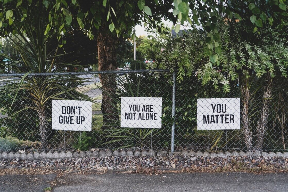 Image of a fence with trees behind and signage saying you are not alone.