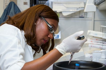 Fêmea cientista em branco com uma pipeta branca sobre um prato de cerâmica. Ela é de pele castanha, usa óculos e está claramente num laboratório.