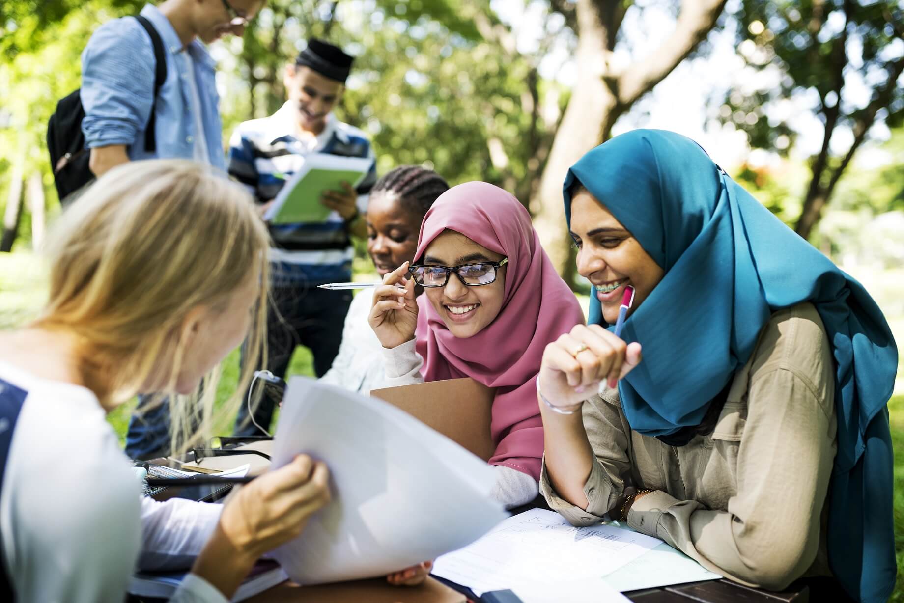 Fotografía de un grupo de jóvenes de la región de Oriente Medio sentados alrededor de una mesa en un parque, interactuando entre sí y mirando material impreso en papel.