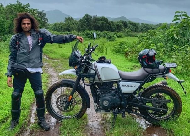 Influencer Zerxes is wearing blue jeans and a denim jacket and is standing next to a motorbike amidst a green landscape.