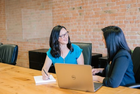 Une femme en chemise sarcelle assise à côté d'une femme en blazer sombre se souriant l'une à l'autre tout en discutant de travail.